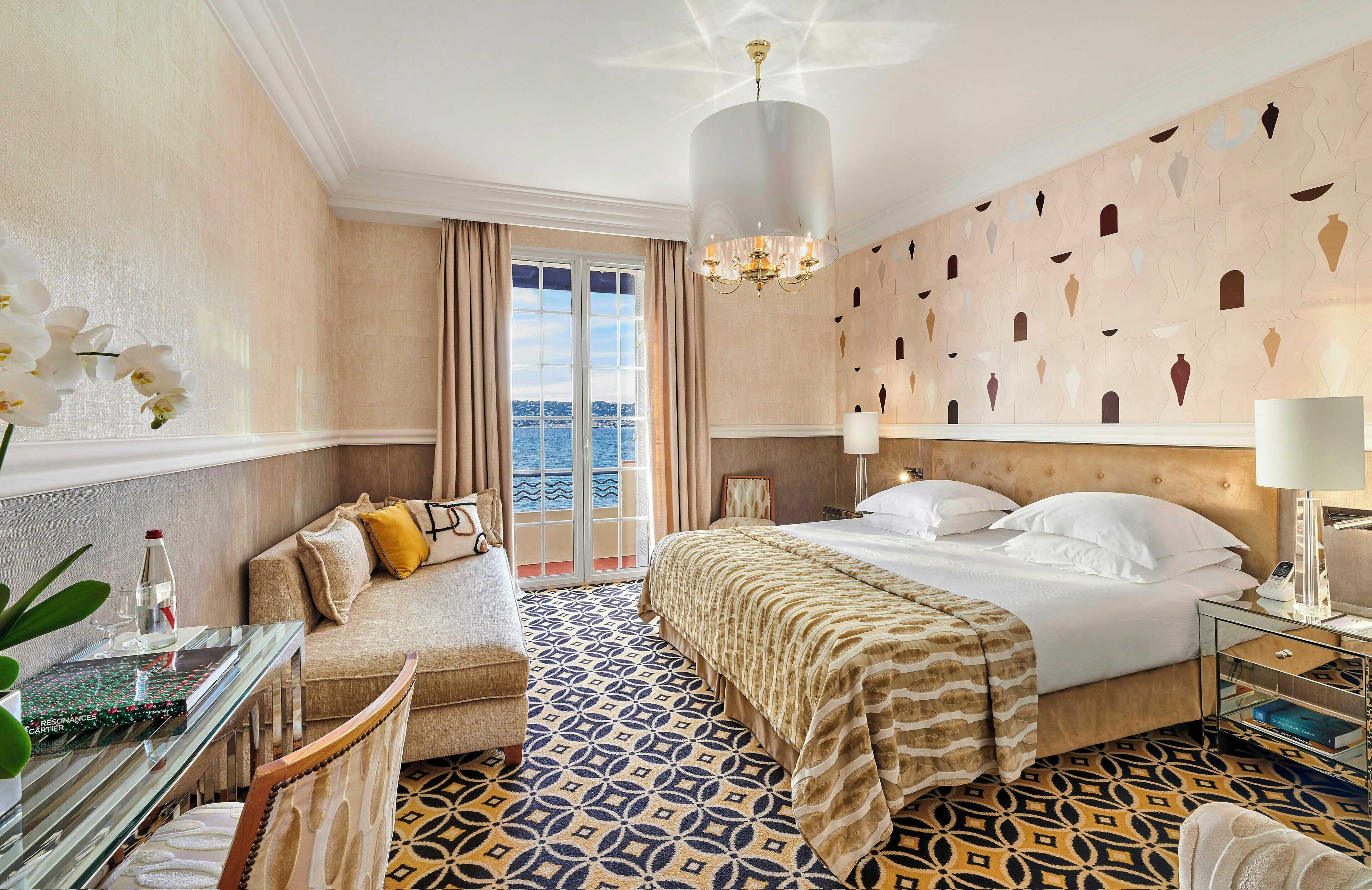 Una camera d'albergo con un letto, un divano, una scrivania con una bottiglia, una sedia, un pavimento a motivi e un lampadario, con vista sul mare dalla finestra.