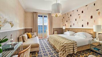 Una camera d'albergo con un letto, un divano, una scrivania con una bottiglia, una sedia, un pavimento a motivi e un lampadario, con vista sul mare dalla finestra.