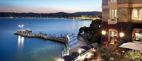 A waterfront restaurant with outdoor seating overlooking a calm body of water, a dock, and a stone building with multiple windows.
