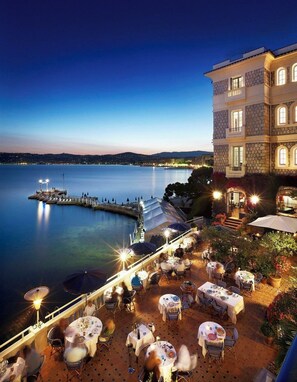 A waterfront restaurant with outdoor seating overlooking a calm body of water, a dock, and a stone building with multiple windows.
