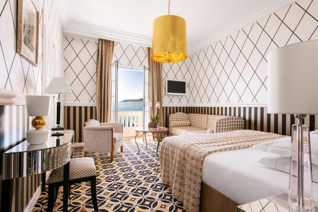 A hotel room with a bed, a desk, a chair, a TV, and a view of the sea.