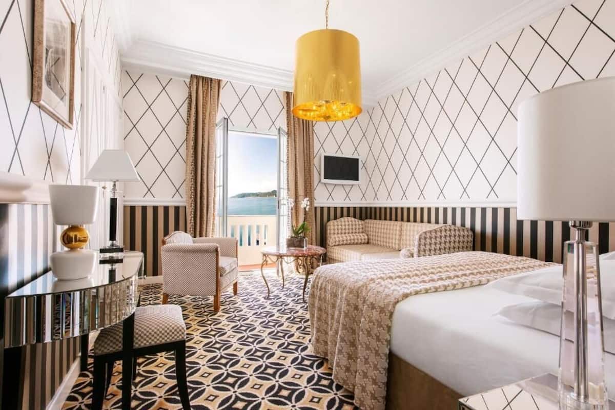 A hotel room with a bed, a desk, a chair, a TV, and a view of the sea.