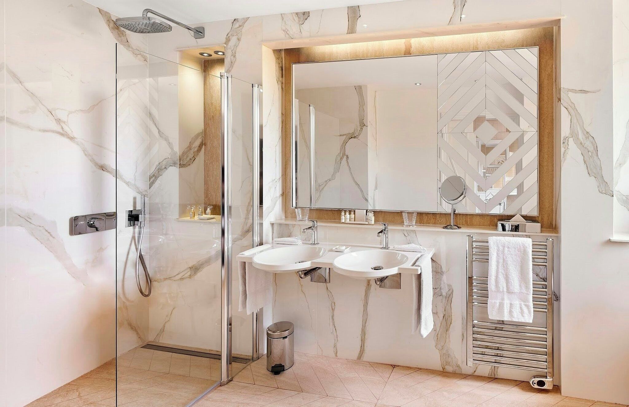 Un baño moderno con ducha de vidrio, dos lavamanos, un espejo grande y un perchero para toallas.