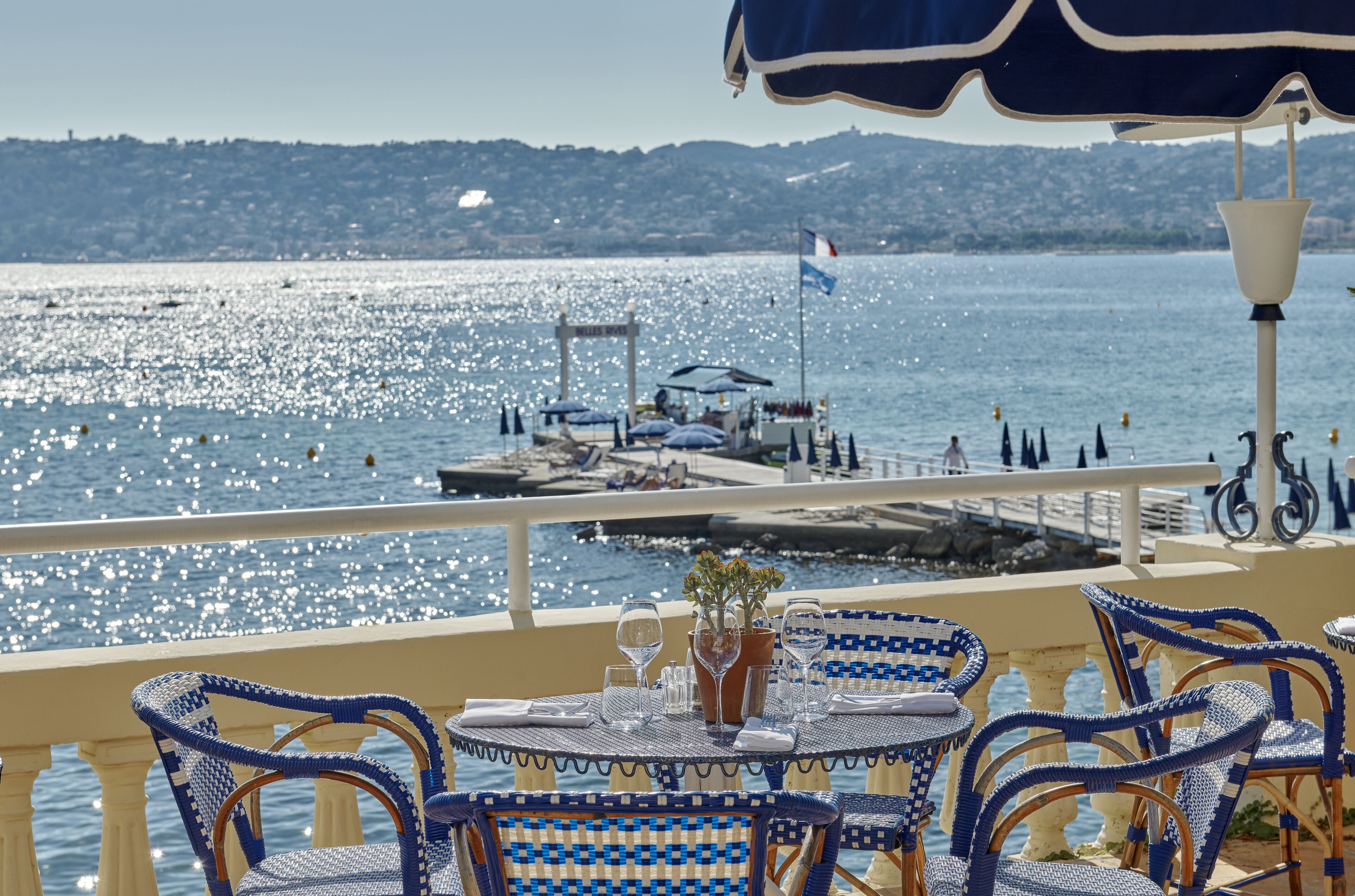Terraza con sillas y mesas de mimbre, con vistas a un cuerpo de agua con barcos y un muelle.