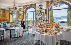 A hotel dining area with tables set for breakfast, a view of the sea, and a woman arranging items on a table.