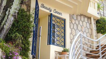 Entrada de un edificio con un letrero que dice 'Beauty Corner', una pared de piedra, un portón metálico y una escalera que sube.