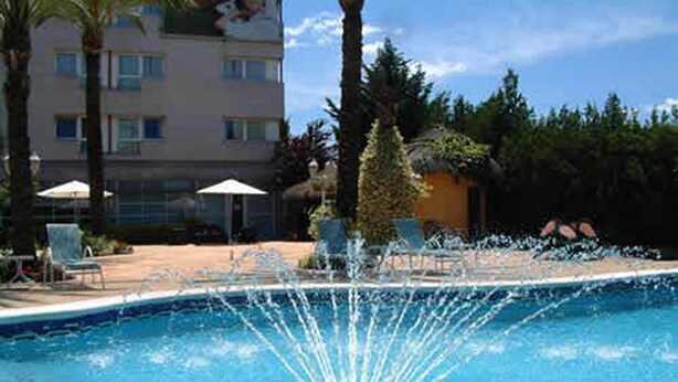 Una piscina con una fuente, rodeada de sillones de playa y sombrillas, con un edificio al fondo.