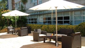 Un patio al aire libre con mobiliario de mimbre, dos sombrillas y vista a un edificio moderno con ventanas amplias.