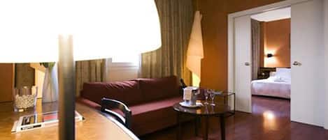 A hotel room with a wooden floor, a red sofa, a small table with a lamp, and a bed in the background.
