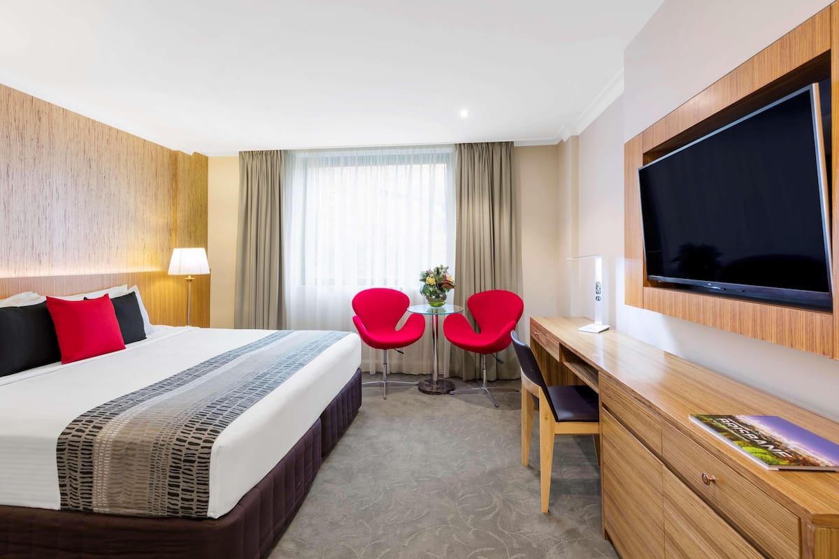 A hotel room with a large bed, a desk with a flat-screen TV, two red chairs, and a small table with a vase.