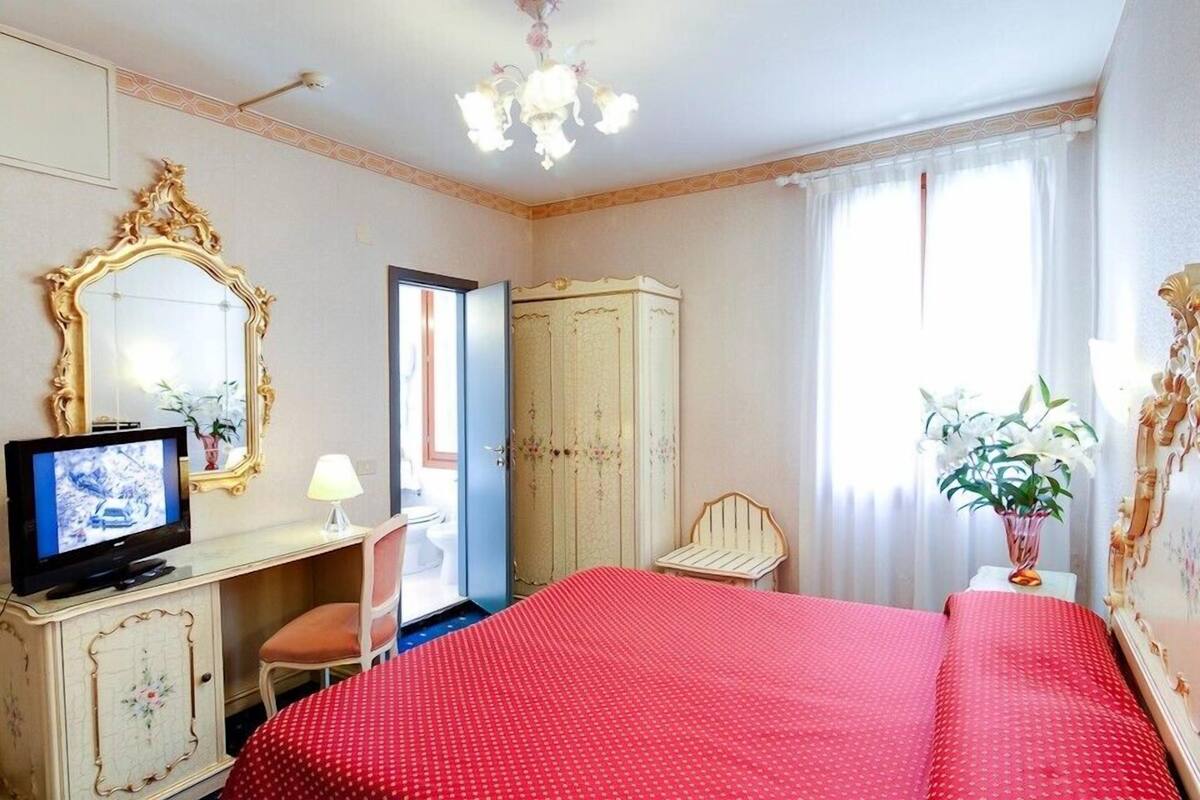 A hotel room with a bed, a television, a desk with a lamp, a wardrobe, a chair, and a window with curtains.