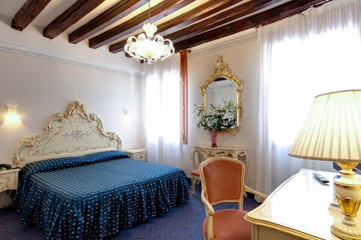 A hotel room with a bed, a desk, a chair, a lamp, a mirror, and a chandelier.
