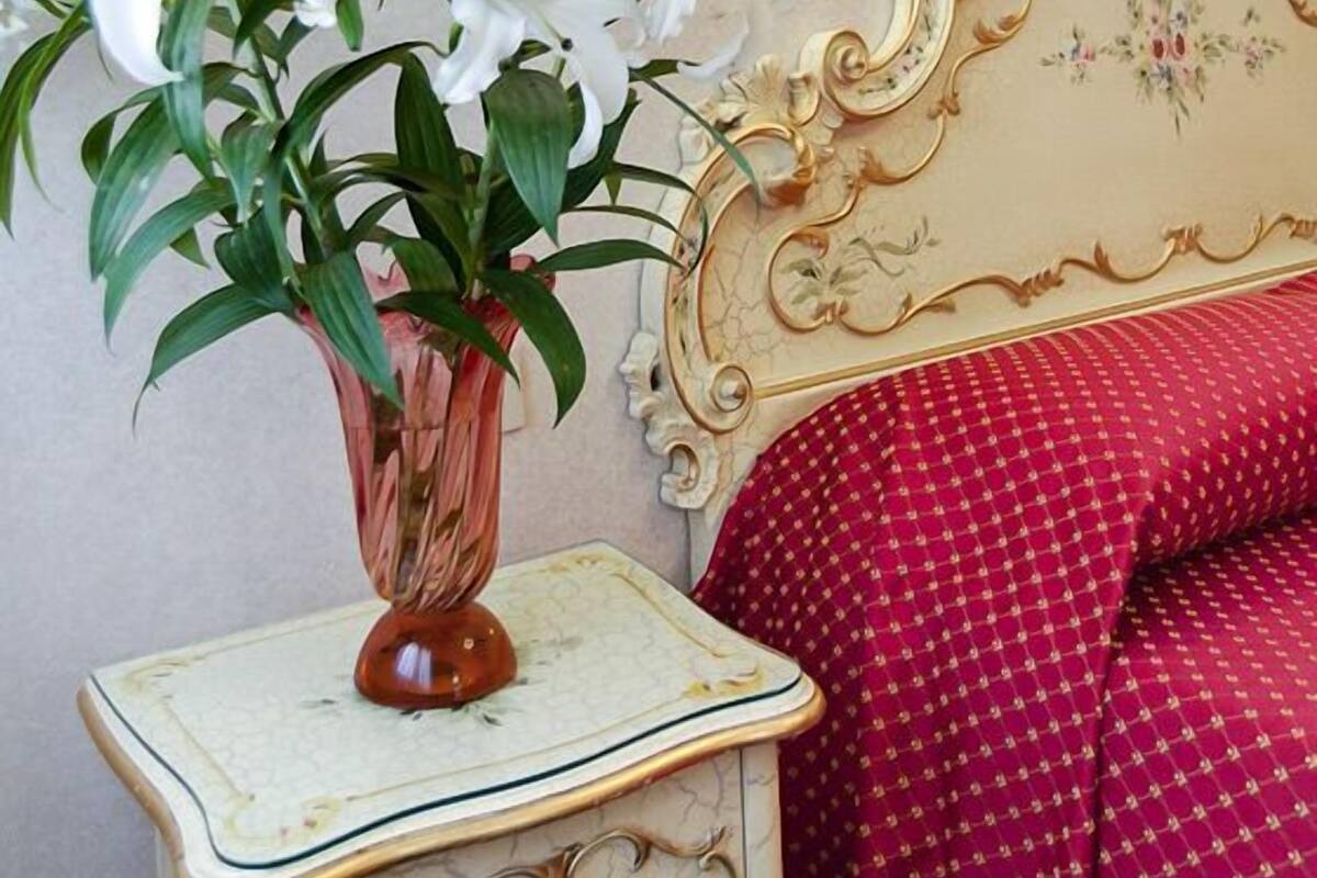 A vintage-style bedside table with a vase of white flowers, a decorative headboard, and a red patterned bedspread.