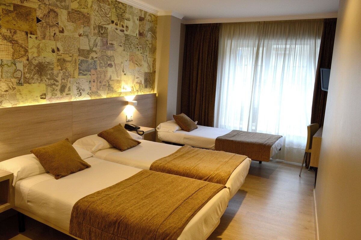 Ein Hotelzimmer mit zwei Betten, einer Wand mit Stadtplänen, einem Schreibtisch und einem großen Fenster mit Vorhängen.