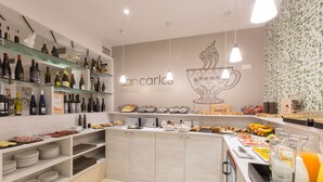 A hotel breakfast area with shelves of wine bottles, a buffet counter with various food items, and a wall with the hotel's name and logo.