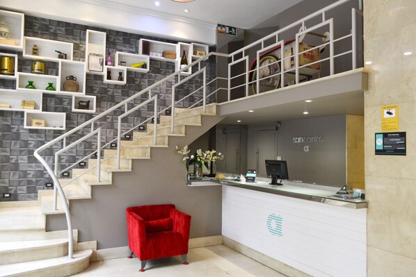 Un espace d’accueil avec un escalier, un fauteuil rouge et des étagères remplies de divers objets.