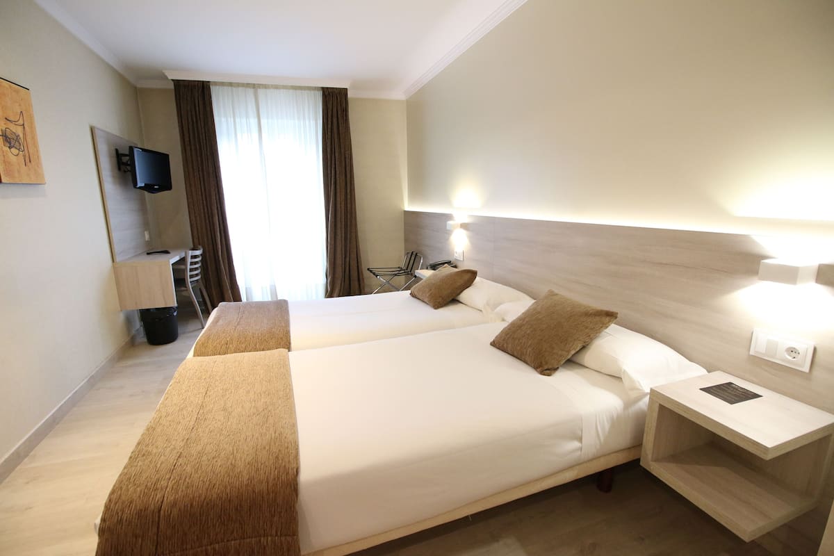 Habitación de hotel con dos camas, un televisor montado en la pared, un escritorio con una silla y una ventana con cortinas.