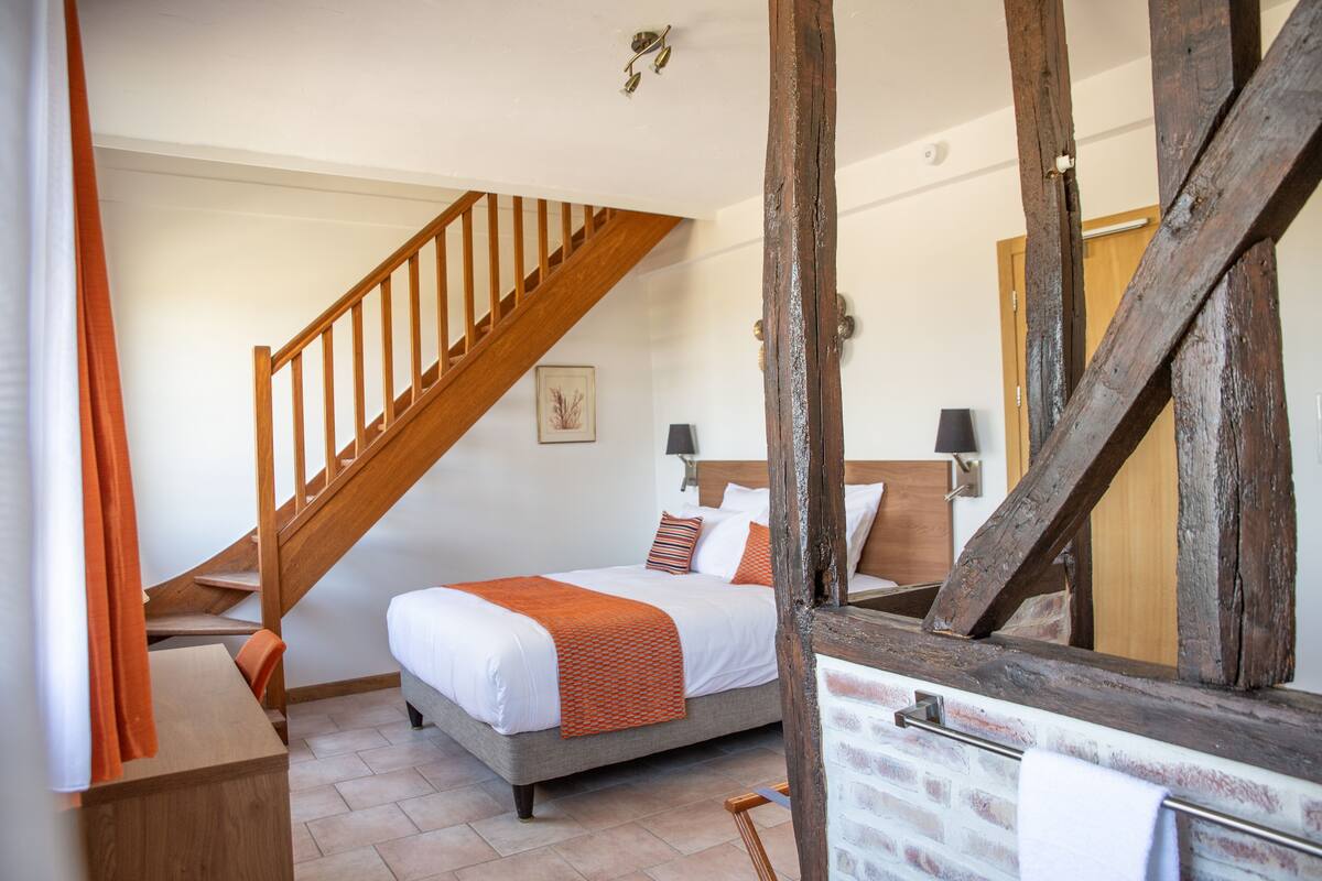Une chambre avec un escalier en bois, un lit avec une literie blanche et des touches de couleur orange, une cheminée en briques et une porte en bois.