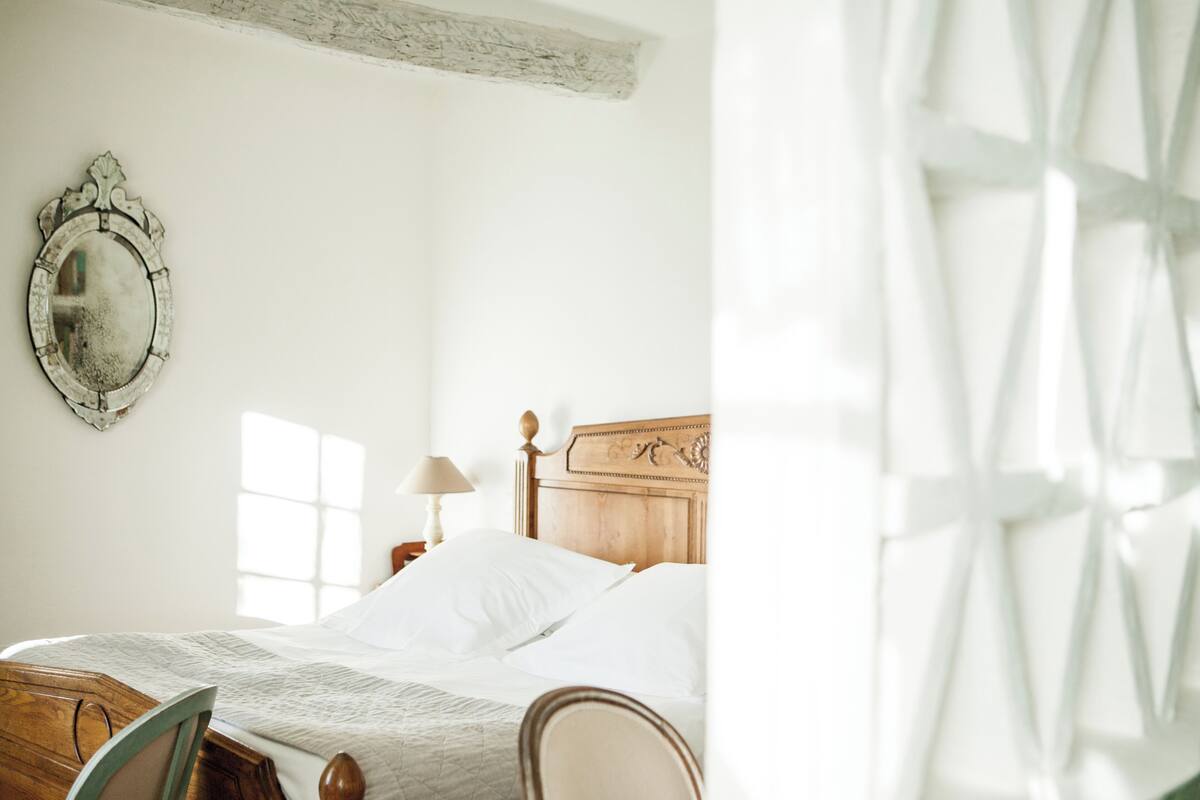 Una cama bien hecha con cabecera de madera, un espejo en la pared y una ventana que deja entrar luz natural.