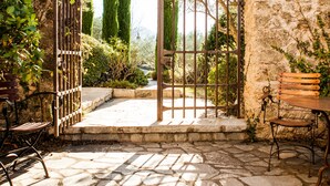 An arched stone gateway leading to a courtyard with a stone floor, surrounded by vegetation and a bench.