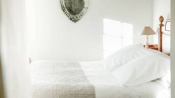 Una cama bien hecha con sábanas blancas, una lámpara de noche y un espejo en la pared.