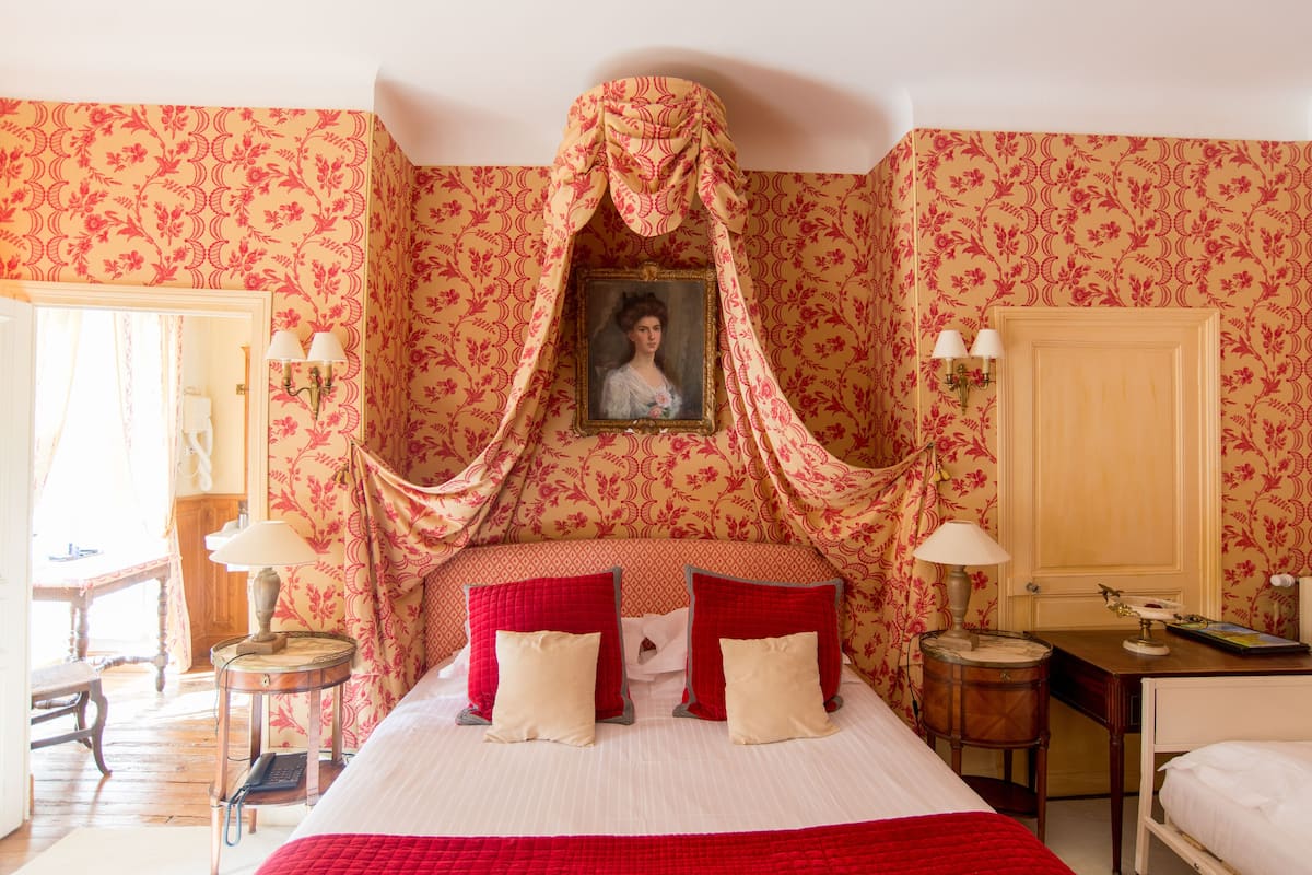 Habitación de hotel con una cama grande, dosel, un retrato en la pared y un escritorio con lámpara.