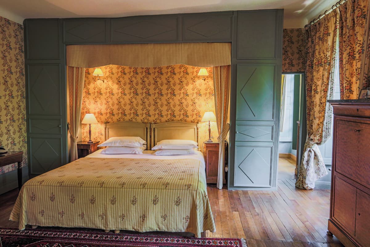 Un dormitorio con una cama grande, dos mesitas de noche con lámparas, papel tapiz floral y un aparador de madera.