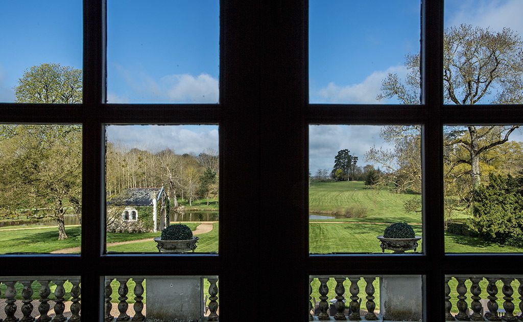 Vista a través de una ventana con barandilla, que muestra un césped cuidado, un edificio pequeño y árboles.