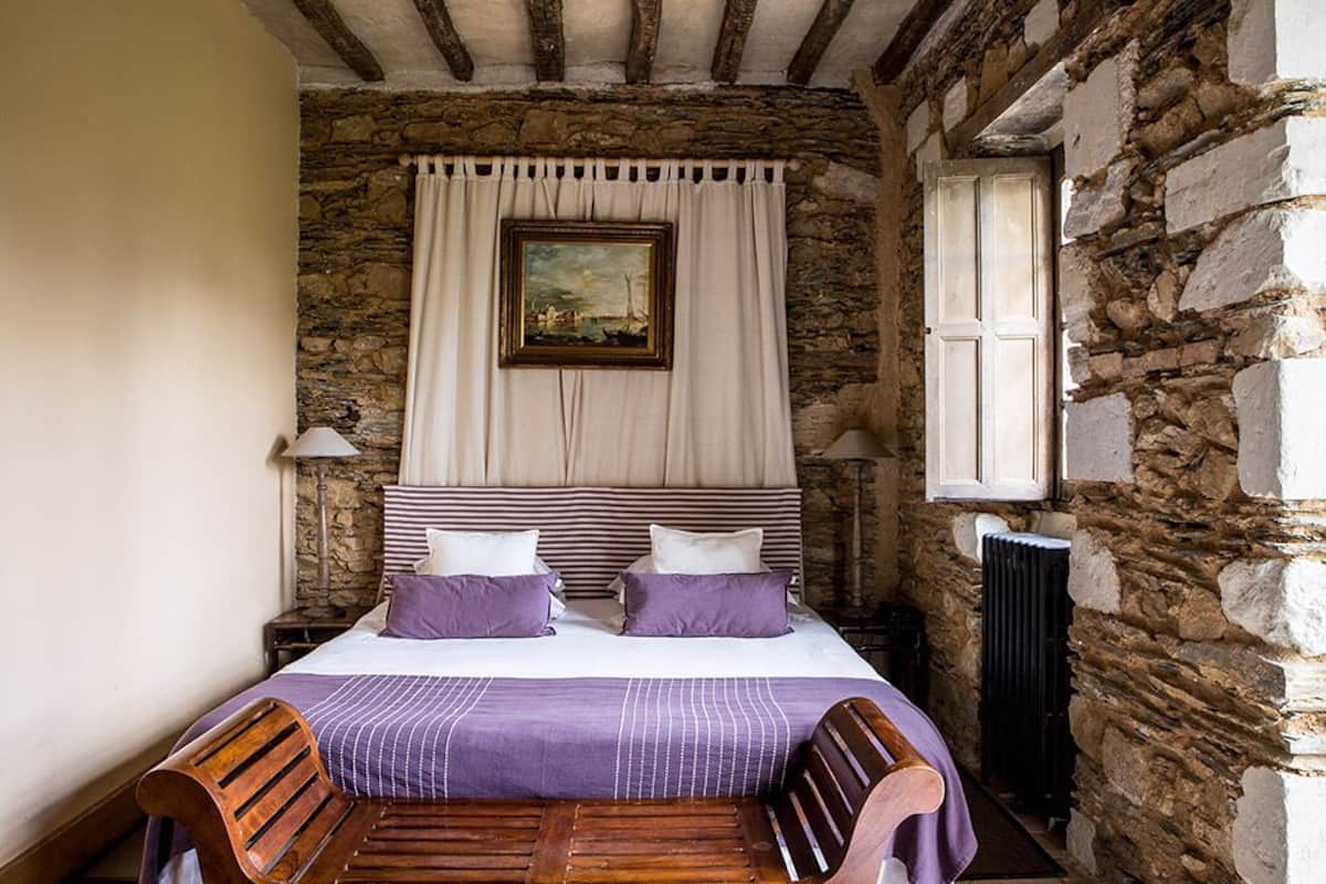 Un dormitorio con pared de piedra, cama con ropa de cama púrpura, cabeceros de madera y un cuadro sobre la cama.