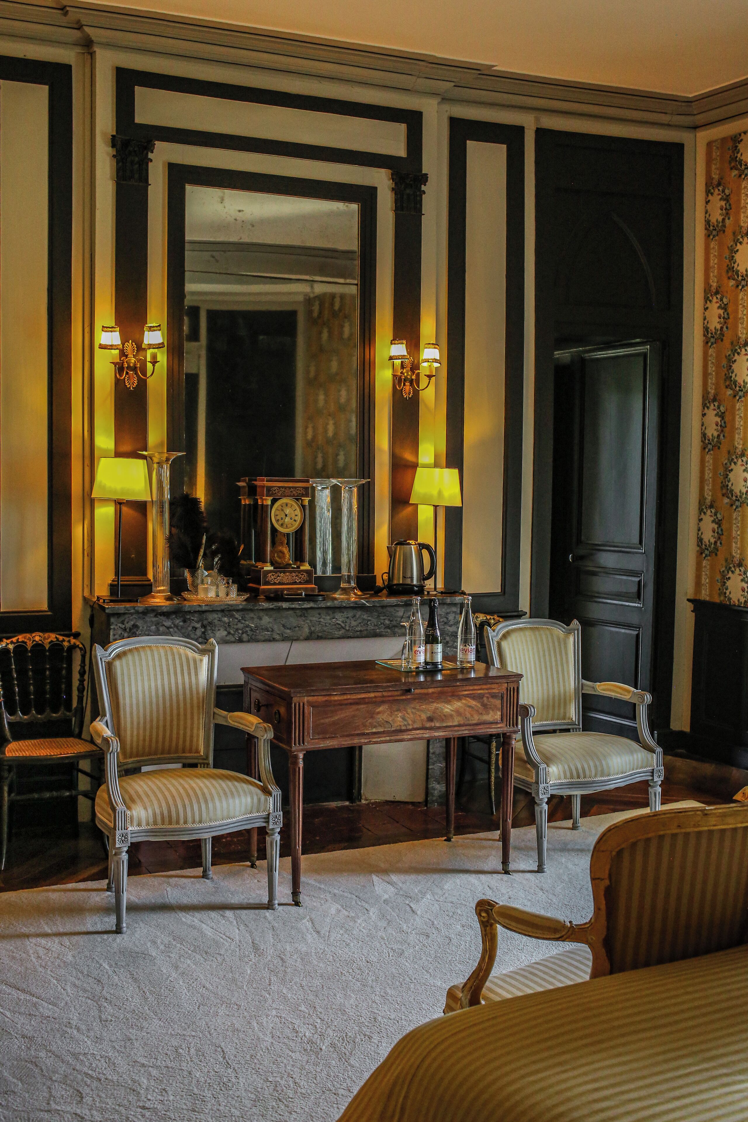 A room with a large mirror, two lamps, a clock, and a coffee pot on a marble countertop. There are four upholstered chairs and a wooden table.