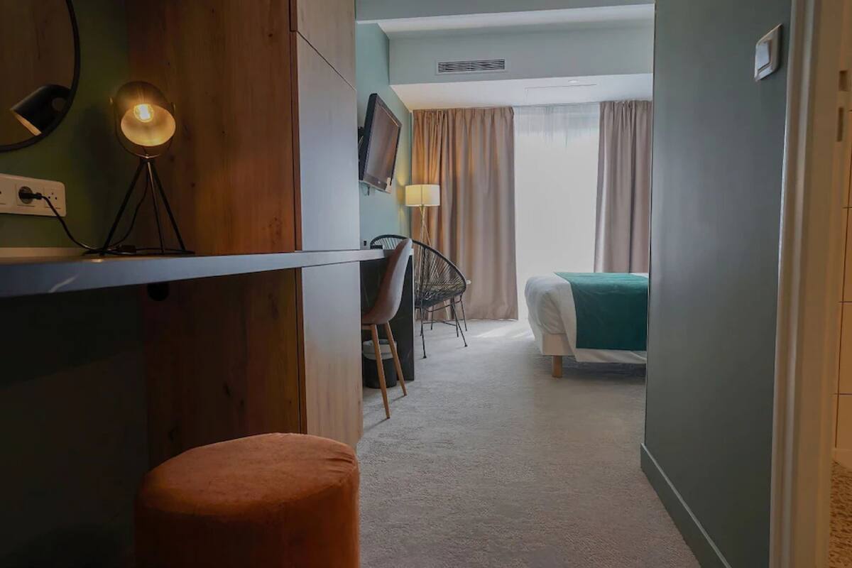 A hotel room with a bed, a desk with a lamp, a chair, a TV, and a window with curtains.