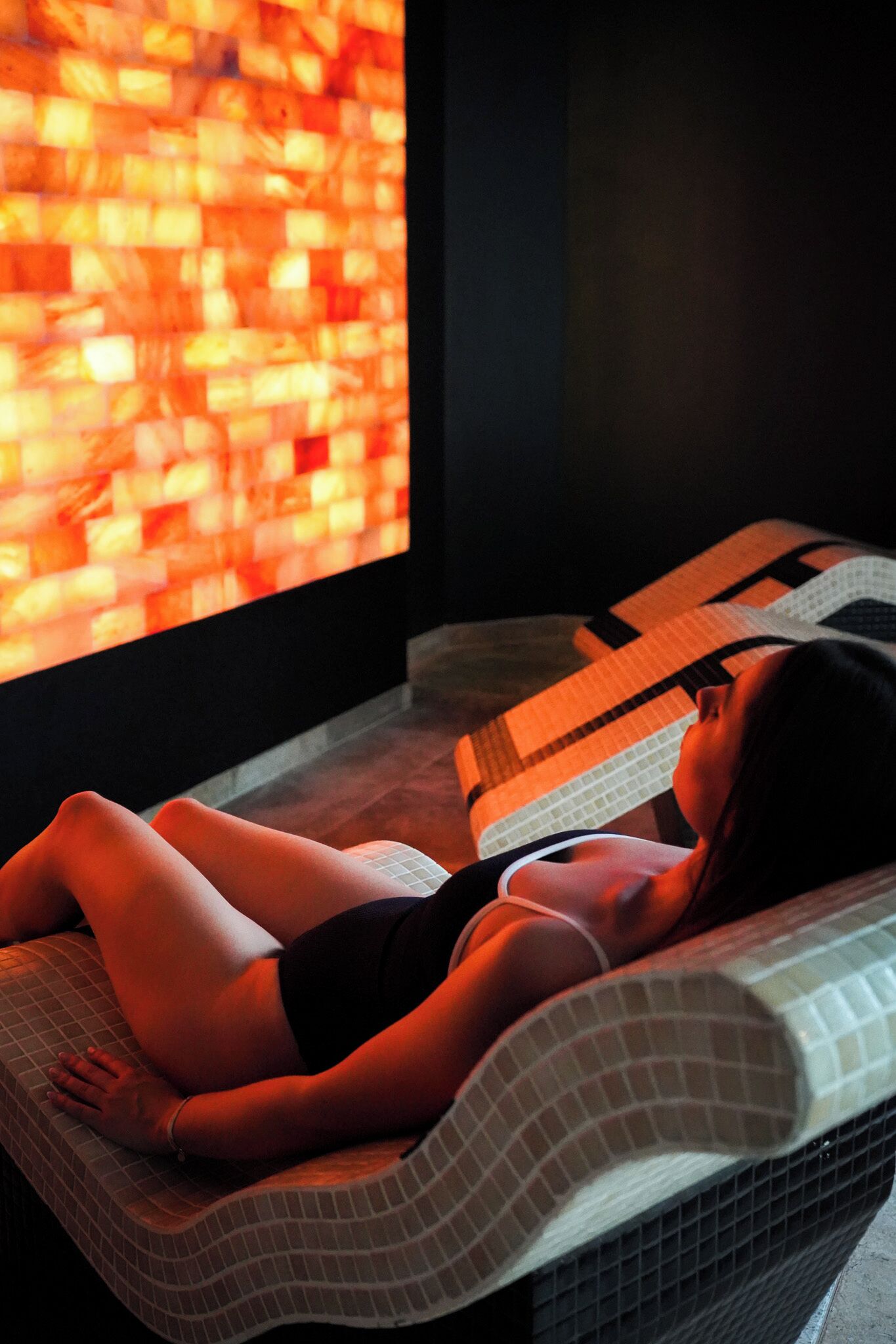 A person lying on a massage table with a patterned cushion, in a room with a large window displaying a mosaic wall.