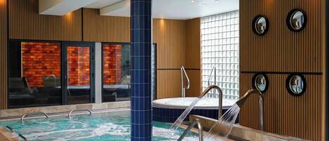 A modern indoor swimming pool with a jacuzzi, wooden paneling, and a glass wall with a brick pattern.