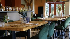 Restaurante com mesas de madeira, cadeiras estofadas verdes e um bar com potes de temperos.