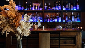 Uma área de bar com balcão de madeira, um vaso com flores secas e prateleiras cheias de garrafas.