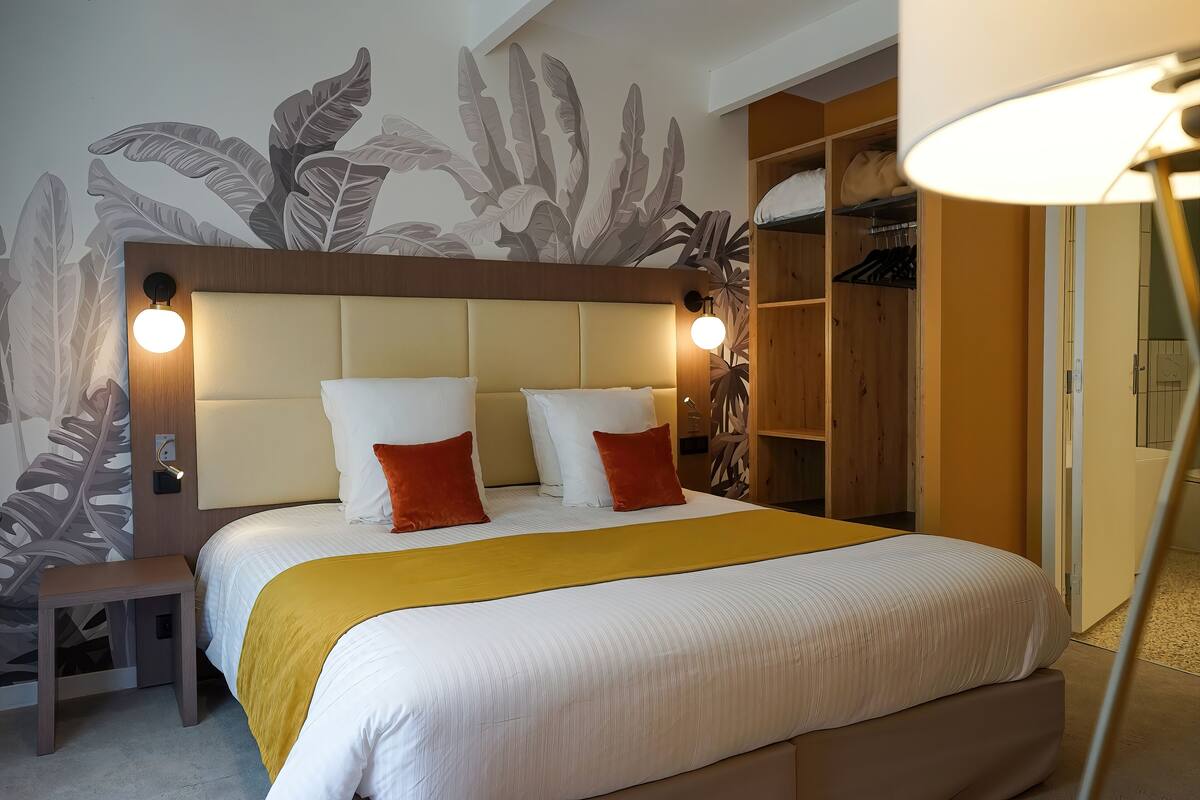 A neatly made bed with a yellow throw, a wooden headboard, and a wall with a leaf pattern.