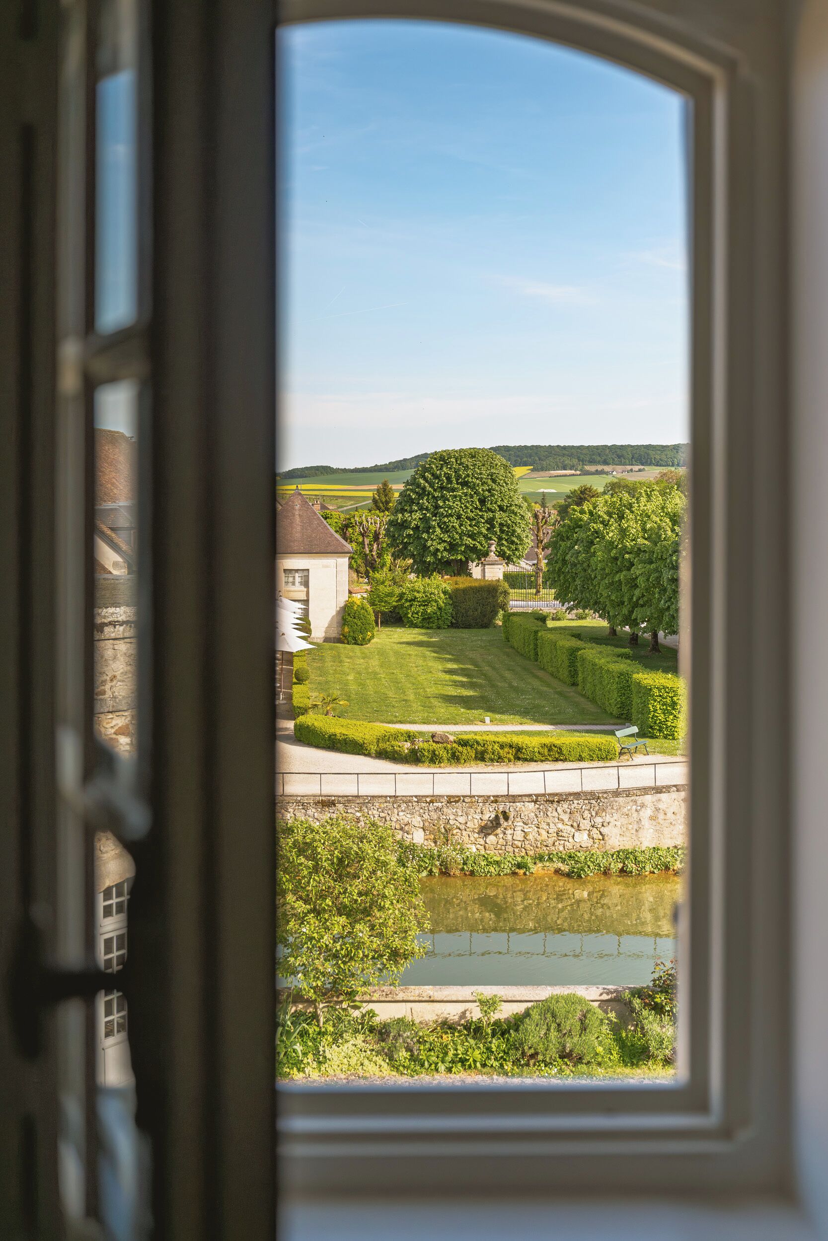 A view through a window of a well-maintained garden with a pond, neatly trimmed hedges, and a building in the background.