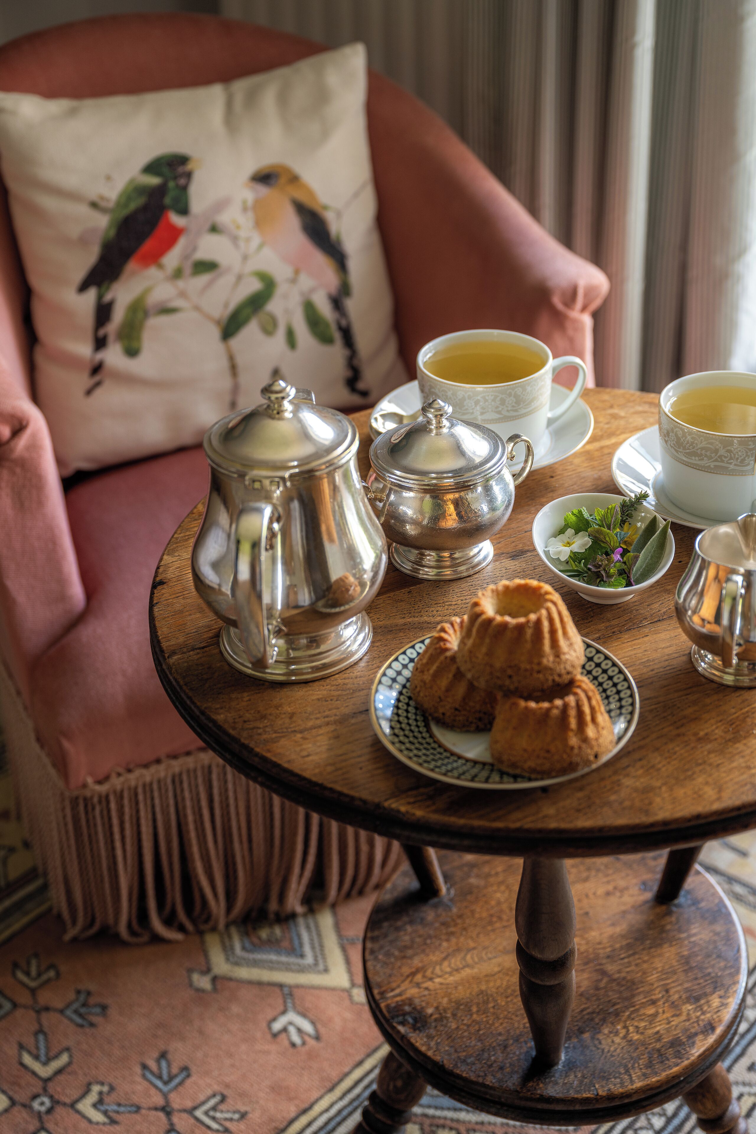 A cozy room with a round wooden table set for tea, a cushioned chair with a decorative pillow, and a patterned rug.