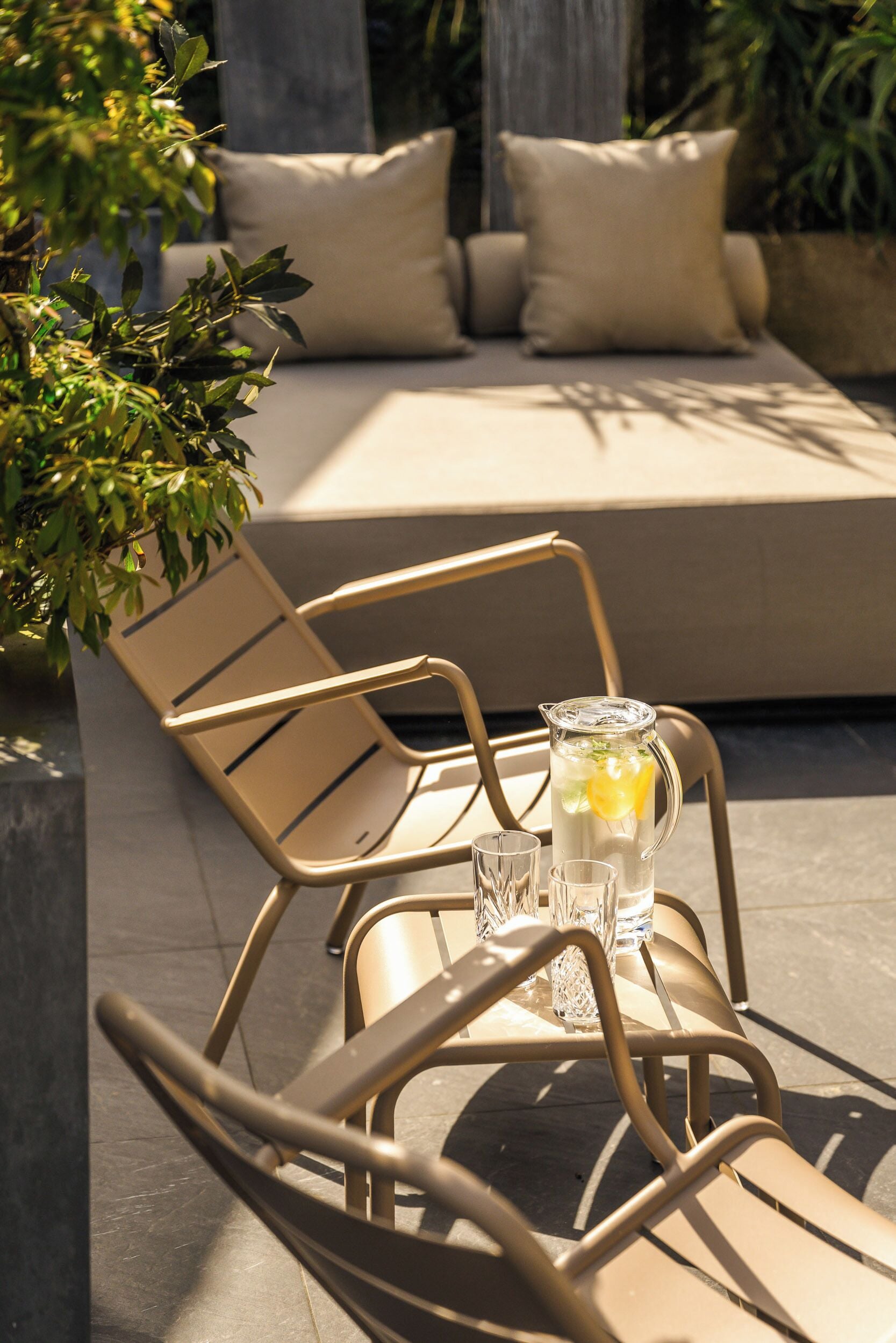 Un espace extérieur ensoleillé, aménagé avec des sièges, comprenant une chaise en métal, une table avec une carafe et des verres, et un canapé avec des coussins.