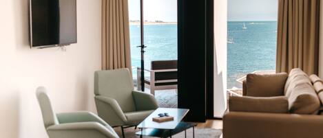 Un salon moderne doté d’un téléviseur, de deux fauteuils, d’un canapé et offrant une vue sur la mer grâce à de grandes fenêtres.