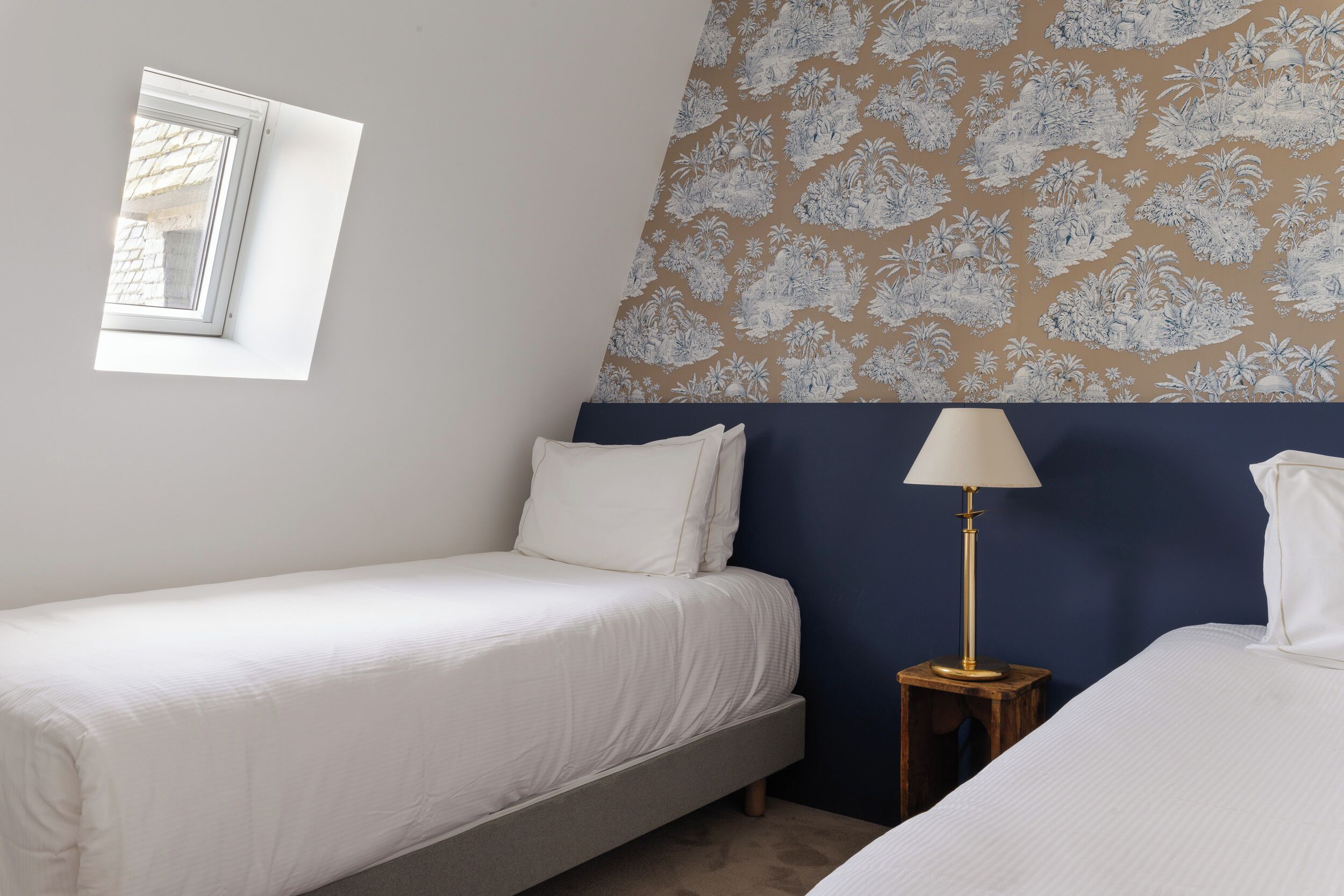 A bedroom with two beds, a window, a wall with a floral pattern, and a bedside lamp.