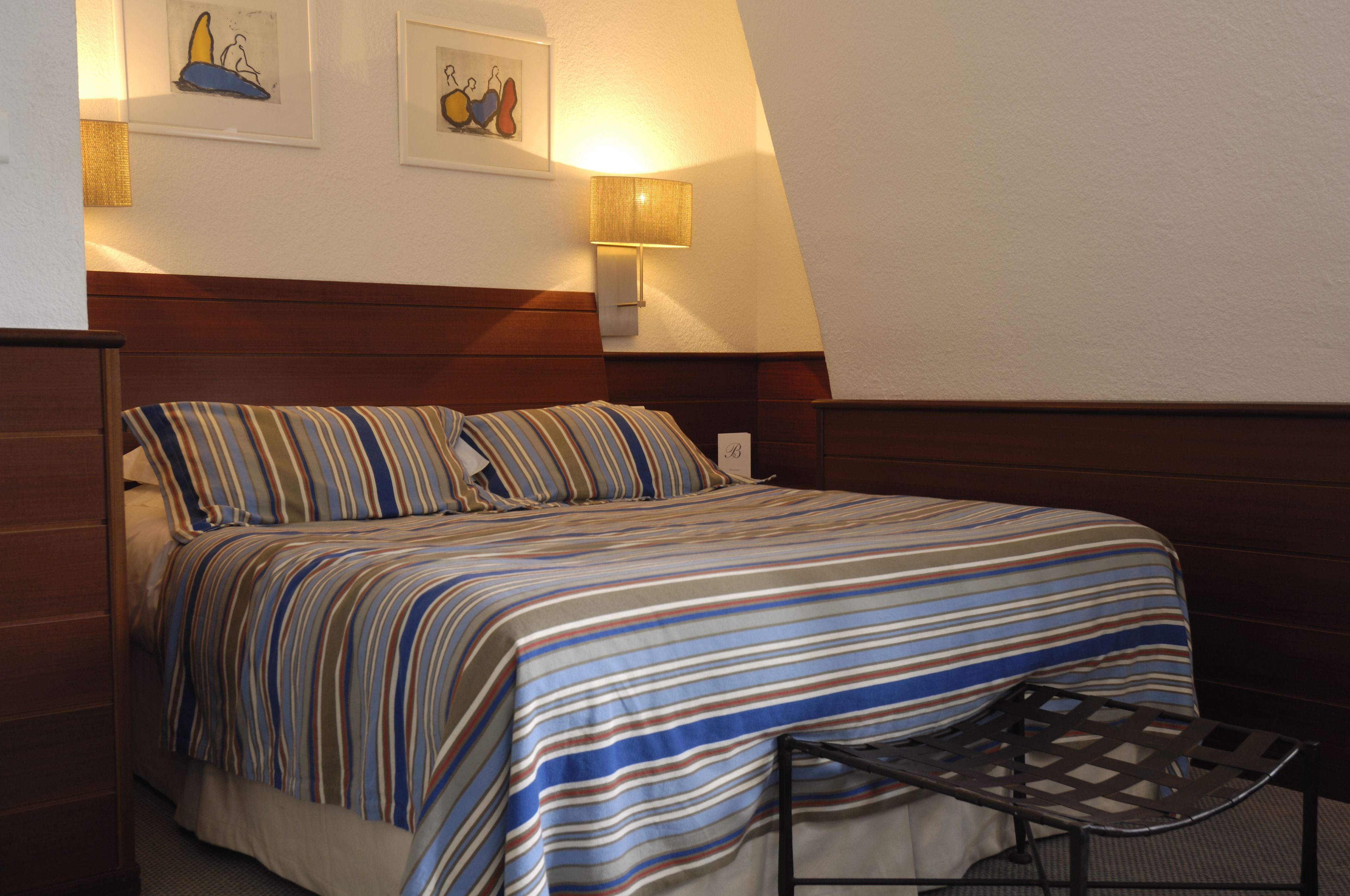 A neatly made bed with striped bedding, a bedside lamp, and two framed artworks on the wall.