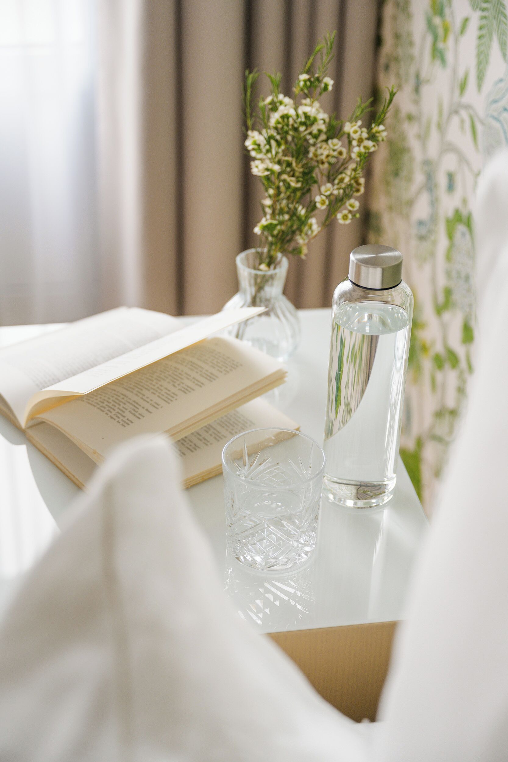 Une table blanche avec un livre, un verre transparent et une bouteille d’eau.
