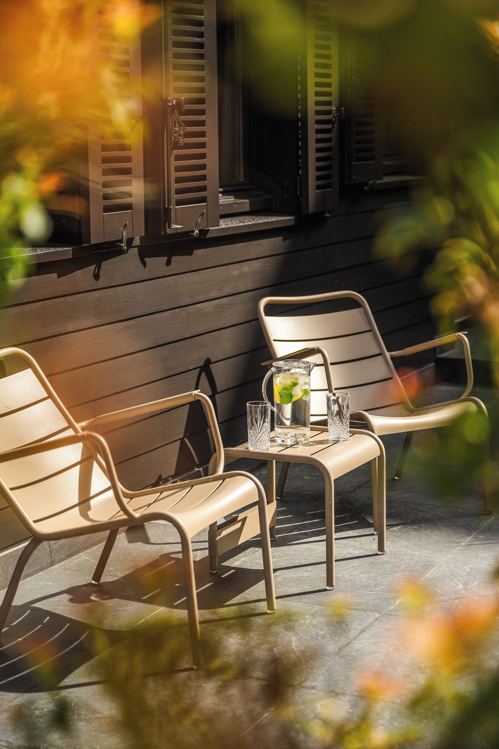 Une terrasse avec deux chaises et une table, une carafe d’eau et un verre.