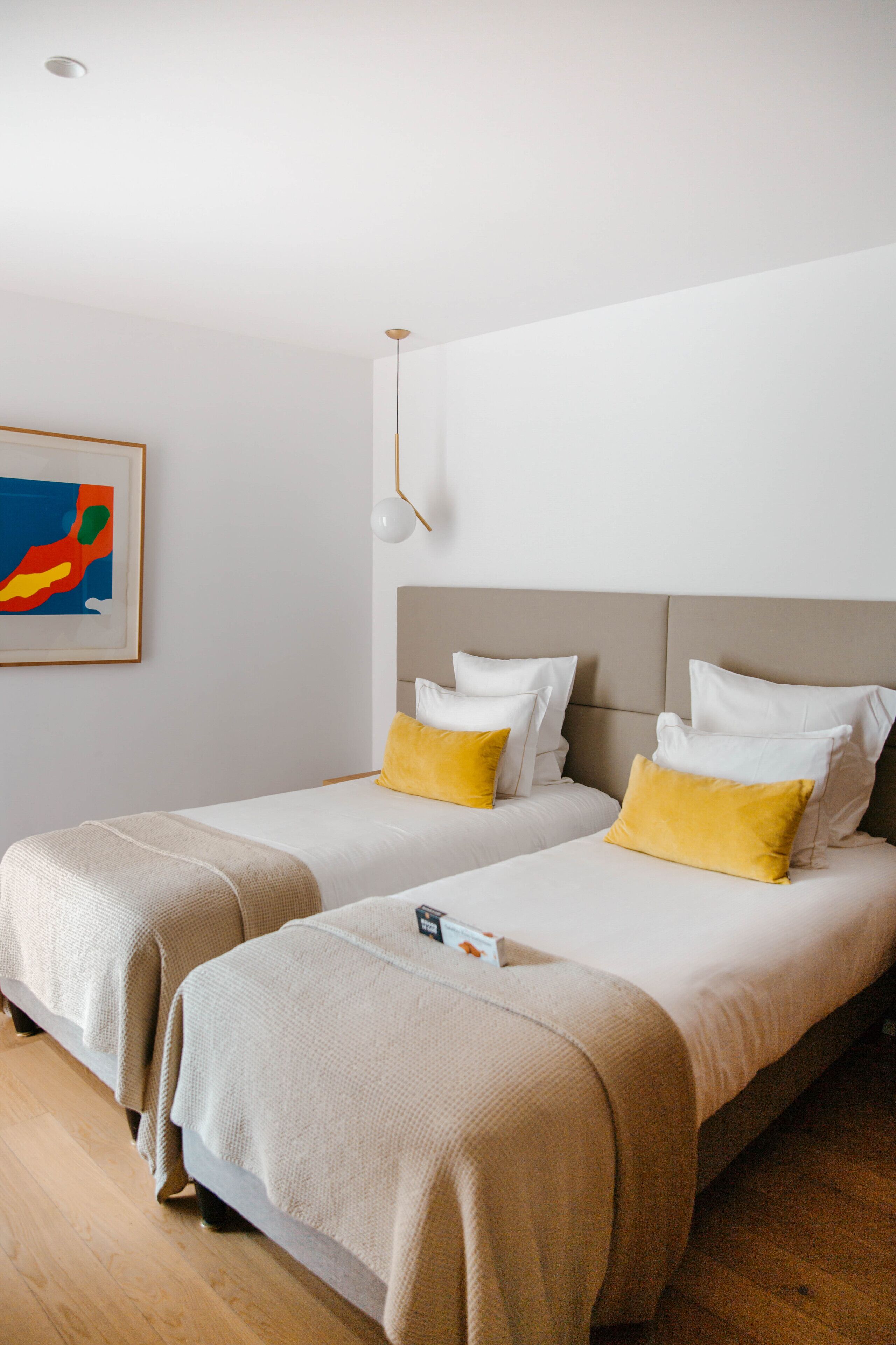 Une chambre d’hôtel avec deux lits, chacun doté d’une tête de lit, de draps blancs et d’oreillers jaunes. Un tableau encadré orne le mur, et une suspension lumineuse se trouve au-dessus des lits.