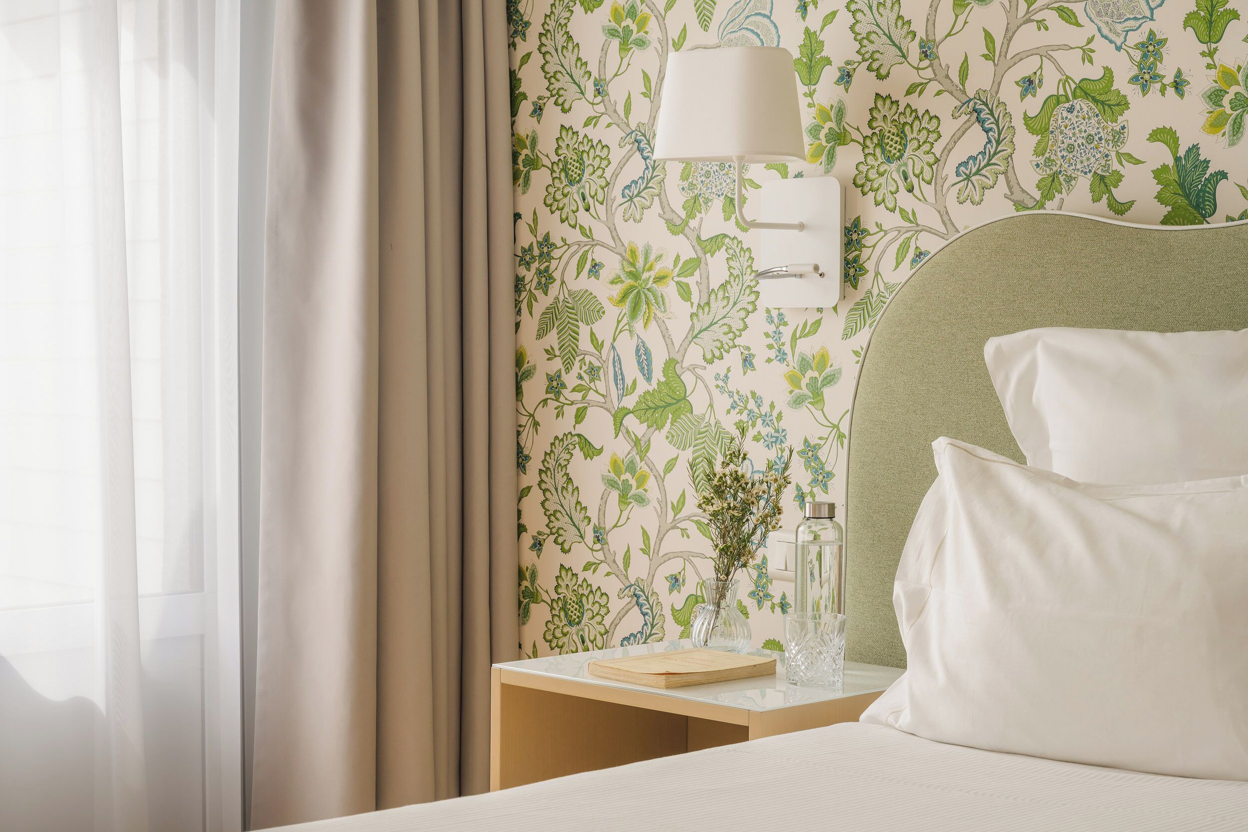 Una cama bien hecha con almohadas blancas, una mesita de noche con lámpara y papel tapiz floral.