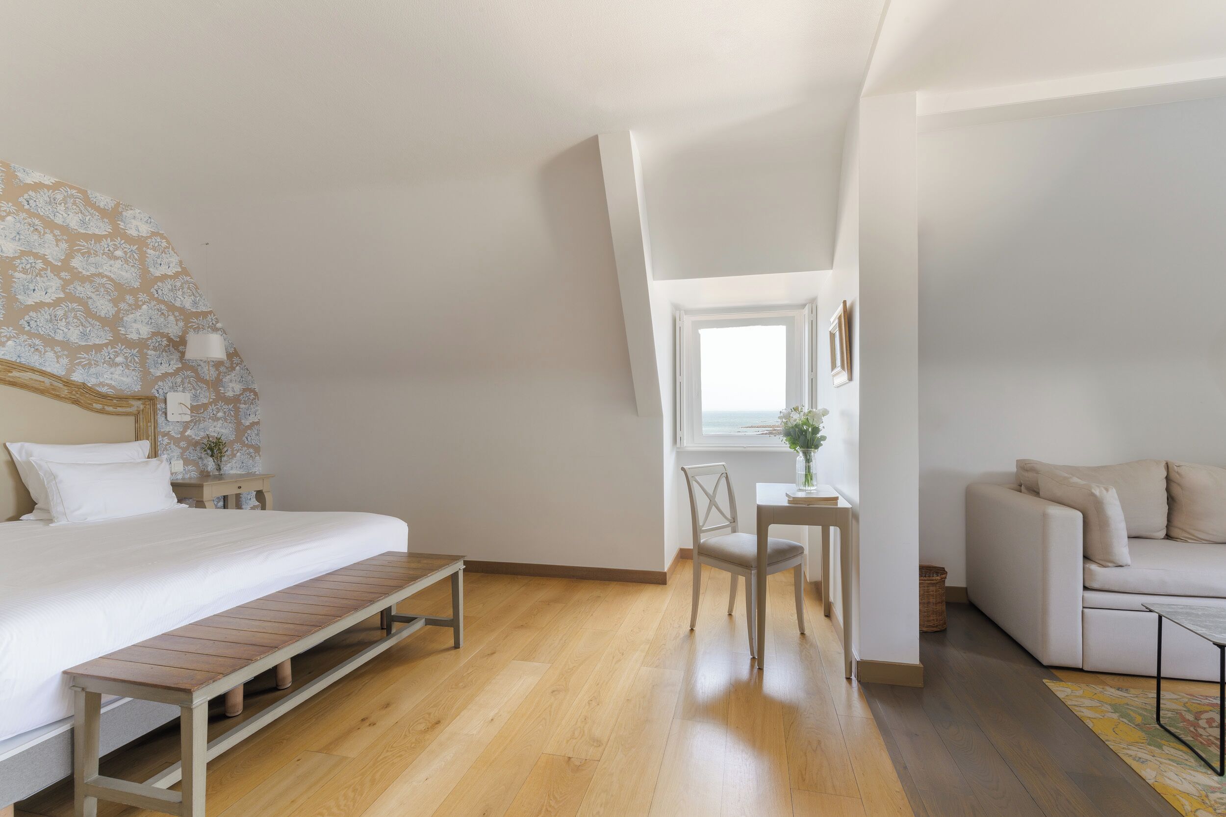Un dormitorio con una cama, una banca, una silla y una mesita de noche con un florero.