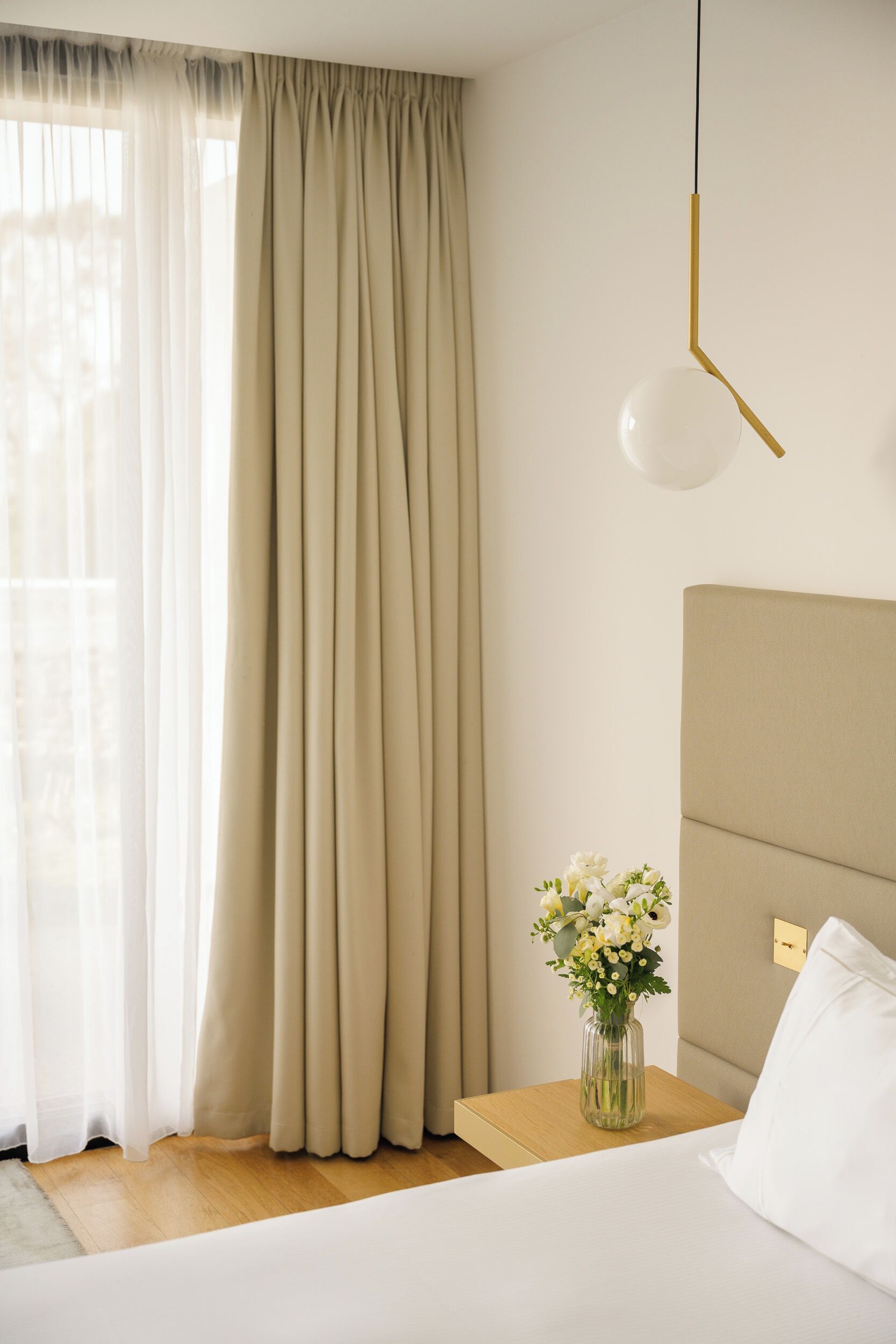A bedroom with a bed, a nightstand, a vase with flowers, a lamp, and curtains.