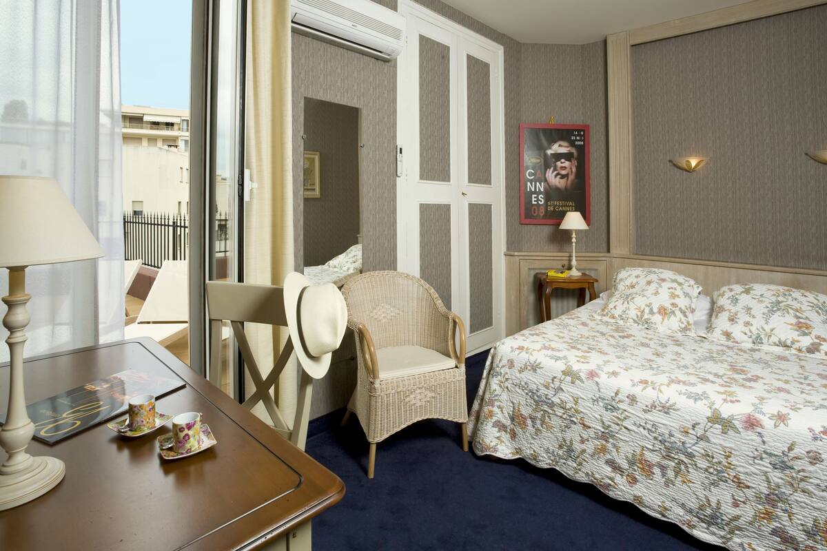 A hotel room with a bed, a wicker chair, a table with a lamp, and a poster on the wall.