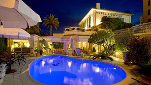 Una piscina con sillas de descanso y sombrillas, rodeada por un hotel llamado HOTEL L'Olivier.